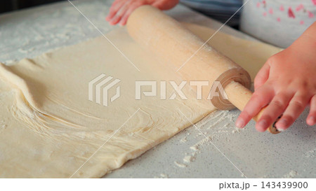 Hands rolling dough with a wooden rolling pin on a floured kitchen counter, preparing homemade pastry or bread Hands rolling dough with a wooden rolling pin on a floured kitchen counter, preparing homemade pastry or bread 143439900