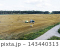 Blue tractor towing baler through harvested wheat field with round straw bales beside gravel farm road under pale sky rural countryside landscape daylight 143440313