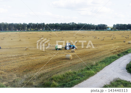 Blue tractor towing baler through harvested wheat field with round straw bales beside gravel farm road under pale sky rural countryside landscape daylight Blue tractor towing baler through harvested wheat field with round straw bales beside gravel farm road under pale sky rural countryside landscape daylight 143440313