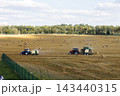 Two wide tractors towing balers across harvested field under soft daylight with distant treeline and fence in foreground, rural agricultural harvest scene 143440315