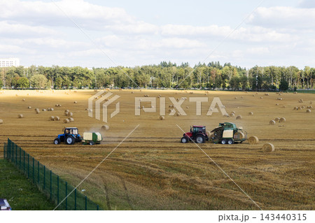 Two wide tractors towing balers across harvested field under soft daylight with distant treeline and fence in foreground, rural agricultural harvest scene Two wide tractors towing balers across harvested field under soft daylight with distant treeline and fence in foreground, rural agricultural harvest scene 143440315