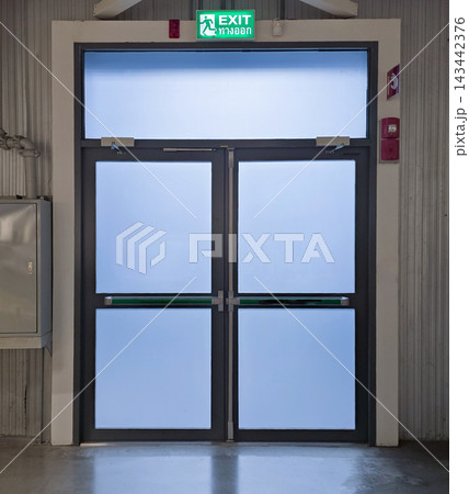Modern double emergency exit doors with frosted glass panels and metal frame. Features an illuminated green exit sign with Thai translation, panic bars, and fire alarm station in a hallway. 143442376