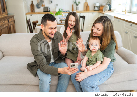 Happy family talk on video call with friends family. Mother father two children holding smartphone talking speaking online. Family mom dad parents baby girls kids smiling laughing enjoying moment Happy family talk on video call with friends family. Mother father two children holding smartphone talking speaking online. Family mom dad parents baby girls kids smiling laughing enjoying moment 143448133