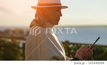Woman in wide brim hat holds smartphone beside calm sea at sunset. Mature traveler gazes toward horizon with thoughtful expression, wrapped in light clothing during peaceful coastal leisure. Woman in wide brim hat holds smartphone beside calm sea at sunset. Mature traveler gazes toward horizon with thoughtful expression, wrapped in light clothing during peaceful coastal leisure. 143448994