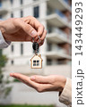 Closeup Hands Landlord Or Property Manager Giving Keys To Tenant Outside Apartment Building. Real Estate Agent Holding The House Key With Home Shaped Keychain To Buyer After Property Purchase 143449293