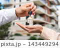 Closeup Hands Landlord Or Property Manager Giving Keys To Tenant Outside Apartment Building. Real Estate Agent Holding House Key With Home Shaped Keychain To Buyer After Property Purchase. 143449294