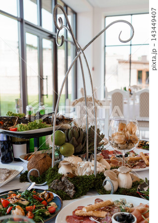 Elegant wedding banquet buffet table with seafood platters, charcuterie, wine glasses, metal arch decor, white chairs, natural light, fresh salad, gourmet spread 143449407