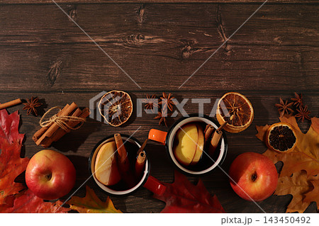 Two colorful enamel mugs with hot autumn drink, mulled wine with apples and autumn fallen leaves, star anise and cinnamon on a wooden table, banner for display, autumn greeting concept, mockup for Two colorful enamel mugs with hot autumn drink, mulled wine with apples and autumn fallen leaves, star anise and cinnamon on a wooden table, banner for display, autumn greeting concept, mockup for 143450492