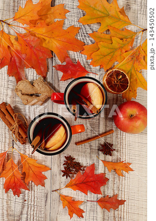 Two colorful enamel mugs with hot autumn drink, mulled wine with apples and autumn fallen leaves, star anise and cinnamon on wooden table, banner for display, autumn greeting concept, mockup for menu Two colorful enamel mugs with hot autumn drink, mulled wine with apples and autumn fallen leaves, star anise and cinnamon on wooden table, banner for display, autumn greeting concept, mockup for menu 143450502