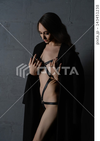 Woman in black leather harness and open coat against dark textured wall, dramatic side lighting, moody atmosphere 143451238