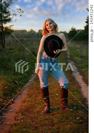 Woman in jeans and tall boots holding a black hat on a rural path Woman in jeans and tall boots holding a black hat on a rural path 143451242