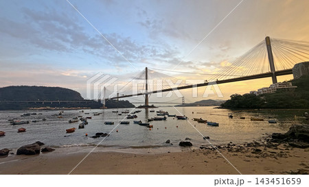 Suspension Hong Kong bridge spanning across a calm bay at sunset with small boats anchored near the shore 143451659