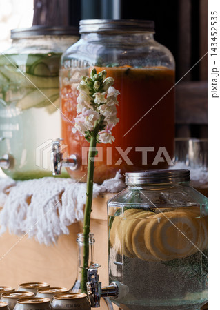 Glass beverage dispensers filled with fruit-infused iced tea lemonade and cucumber water beside a floral sprig at an outdoor event Glass beverage dispensers filled with fruit-infused iced tea lemonade and cucumber water beside a floral sprig at an outdoor event 143452535