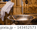 Silver baptismal font with three lit candles and white lace cloth in ornate golden church interior 143452573