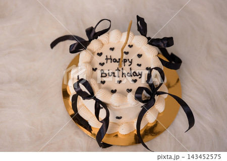 White birthday cake with black ribbons and candles on a gold plate, elegant party dessert with happy message in hearts. White birthday cake with black ribbons and candles on a gold plate, elegant party dessert with happy message in hearts. 143452575