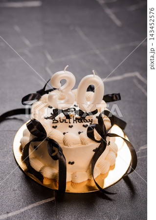 White frosting thirty birthday cake with black ribbons and candles on gold plate against dark grey slate background surface White frosting thirty birthday cake with black ribbons and candles on gold plate against dark grey slate background surface 143452576