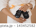 Pregnant woman in white suit holding black baby shoes with bow, maternity concept, gold jewelry, soft studio light. 143452582