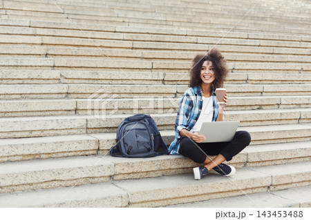 A woman sits on the steps with a laptop on her lap and a coffee cup in her hand. She smiles while looking at the screen. A backpack rests beside her. It is daytime at a busy spot. 143453488