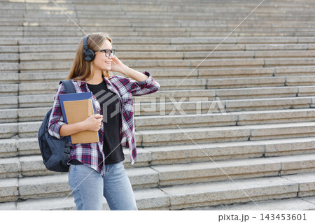 A young person stands on a set of stairs wearing headphones and smiling. They hold a notebook in one hand and have a backpack on their shoulder. 143453601