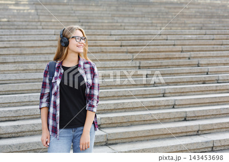A woman with long hair wears headphones and smiles while standing on stone steps. She is dressed casually in a plaid shirt and jeans, looking happy as she enjoys listening to music. 143453698