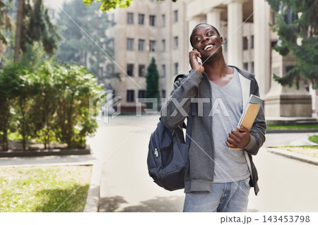 A young man stands outside on campus talking on his phone. He wears a casual outfit and holds books in one hand while carrying a backpack. Trees and buildings are visible in the background. 143453798