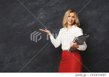 Confident businesswoman pointing away. Woman gesturing with her hand aside and holding notebook, gray studio background, copy space Confident businesswoman pointing away. Woman gesturing with her hand aside and holding notebook, gray studio background, copy space 143453897