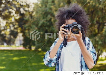 A woman stands in a park holding a camera and looking through the lens. Trees surround her while she prepares to take a photo. Sunlight shines through the leaves. A woman stands in a park holding a camera and looking through the lens. Trees surround her while she prepares to take a photo. Sunlight shines through the leaves. 143453954