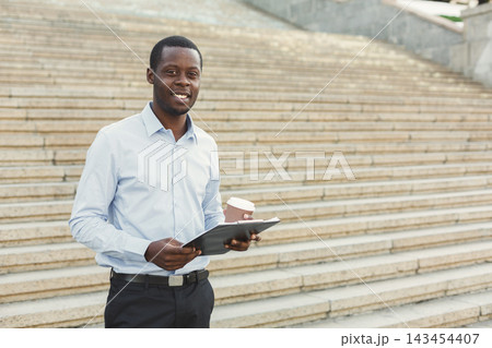A man stands on a set of stone steps in an urban area. He holds a tablet in one hand and a coffee cup in the other. The scene shows a busy and active environment. A man stands on a set of stone steps in an urban area. He holds a tablet in one hand and a coffee cup in the other. The scene shows a busy and active environment. 143454407