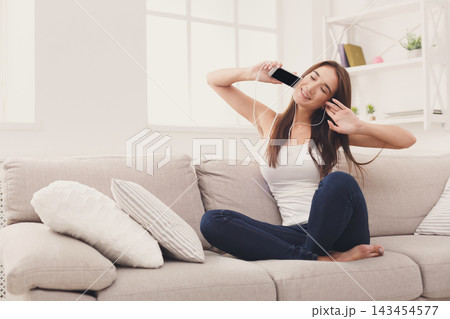 A woman sits comfortably on a couch in her living room, smiling and listening to music on her phone. She enjoys her time alone and feels relaxed. Sunlight fills the space through the windows. 143454577