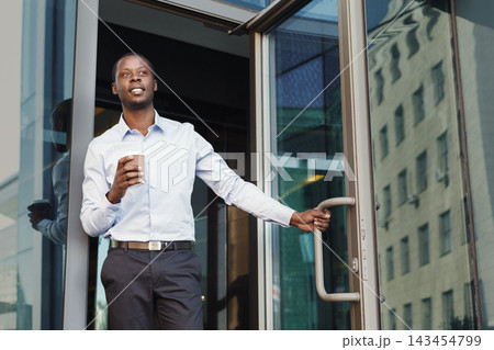 A person holds a coffee cup while standing at the entrance of a modern building. The sun shines on the glass facade, reflecting the surroundings. The location seems busy with activity. 143454799