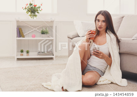 A person sits on the floor with a soft blanket wrapped around them. They hold a drink in one hand and a smartphone in the other. The setting is a light-filled living room with a cozy atmosphere. 143454859