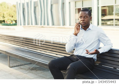A man sits on a black bench in an outdoor area. He talks on the phone while gesturing with his free hand. The background shows a modern building and greenery. It is daytime. 143455443