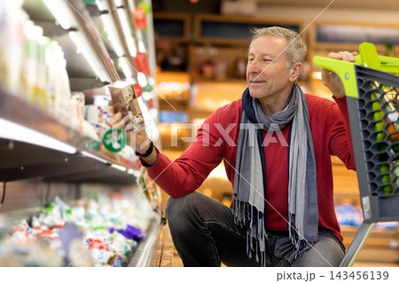 Smiling Elderly Caucasian Man Shopping In Supermarket, Holding Healthy Food Items While Kneeling Down In Aisle, Enjoying His Time Picking Fresh Products In Retail Environment. Business Concept. Smiling Elderly Caucasian Man Shopping In Supermarket, Holding Healthy Food Items While Kneeling Down In Aisle, Enjoying His Time Picking Fresh Products In Retail Environment. Business Concept. 143456139