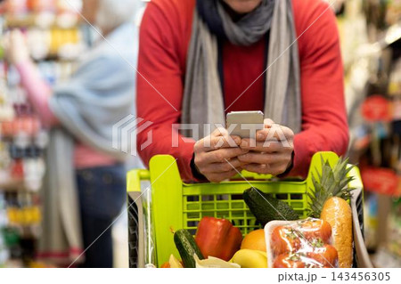 Focused Unrecognizable Person Holding Smartphone In Supermarket Cart Filled With Fresh Groceries, Surrounded By Various Food Products In A Busy Grocery Store Setting. Shopping Concept. 143456305
