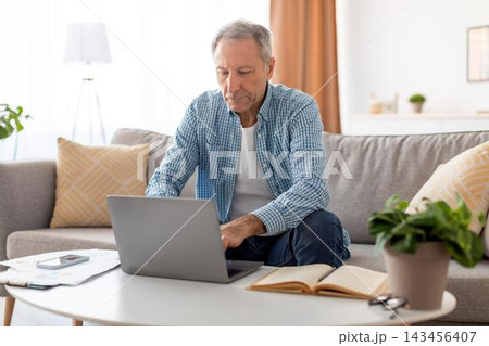 Focused Senior Male Working At Home Office On Laptop, Sitting In Comfortable Position With A Book Nearby, Enjoying Productive Day, In Cozy Modern Living Space. Business Concept. 143456407