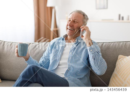 Happy Elderly Caucasian Man Smiling While Conversing On Phone And Holding A Cup Of Coffee, Sitting Comfortably At Home On Sofa. Enjoying Relaxing Moment And Social Connection, Lifestyle Concept. Happy Elderly Caucasian Man Smiling While Conversing On Phone And Holding A Cup Of Coffee, Sitting Comfortably At Home On Sofa. Enjoying Relaxing Moment And Social Connection, Lifestyle Concept. 143456439