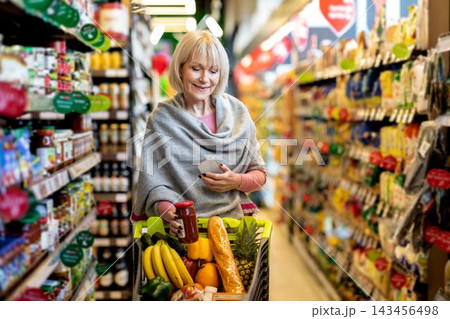 Smiling Elderly Caucasian Woman Shopping In Supermarket, Holding Phone And Choosing Fresh Produce From A Cart While Enjoying Shopping Experience In Store. Weight Loss Concept. 143456498