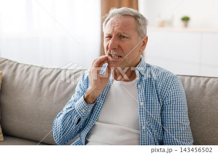Worried Elderly Caucasian Man Holding His Jaw In Pain While Sitting On Couch At Home, Experiencing Dental Discomfort. Indoor Setting Highlighting His Condition And Need For Care, Healthcare Concept. Worried Elderly Caucasian Man Holding His Jaw In Pain While Sitting On Couch At Home, Experiencing Dental Discomfort. Indoor Setting Highlighting His Condition And Need For Care, Healthcare Concept. 143456502