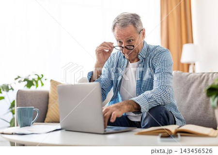 Focused Elderly Caucasian Man Sitting On Sofa Using Laptop For Work Or Leisure At Home, Engaged In Task, Natural Light Coming From Window, Modern Workspace Setting, Business Concept. Focused Elderly Caucasian Man Sitting On Sofa Using Laptop For Work Or Leisure At Home, Engaged In Task, Natural Light Coming From Window, Modern Workspace Setting, Business Concept. 143456526