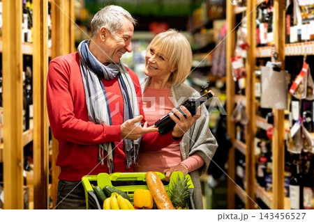 Smiling Elderly Caucasian Man And Woman Enjoying Shopping Together At Supermarket, Holding A Bottle Of Wine In A Cart While Choosing Fresh Produce, Happy Moments In Grocery Store. 143456527