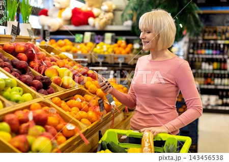 Smiling Caucasian Woman Standing In Grocery Store, Shopping For Fresh Fruits While Holding Smartphone And Basket, Focused On Healthy Eating Choices In Vibrant Setting. Grocery Concept. Smiling Caucasian Woman Standing In Grocery Store, Shopping For Fresh Fruits While Holding Smartphone And Basket, Focused On Healthy Eating Choices In Vibrant Setting. Grocery Concept. 143456538