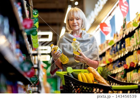 Focused Elderly Caucasian Woman Comparing Two Bottles Of Vegetable Oil While Shopping In Supermarket Aisle. She Is Making Healthy Choices For Her Nutrition. Weight Loss Concept. Focused Elderly Caucasian Woman Comparing Two Bottles Of Vegetable Oil While Shopping In Supermarket Aisle. She Is Making Healthy Choices For Her Nutrition. Weight Loss Concept. 143456539