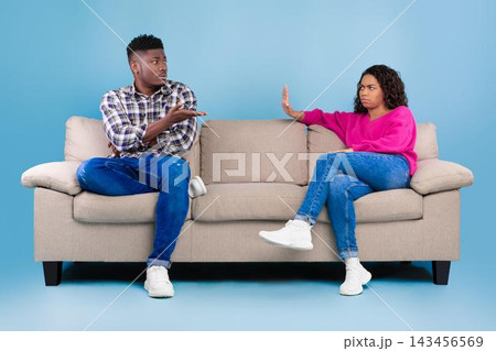 Stressed African American Couple In Their 20s Having Argument While Sitting On Couch At Home. Displaying Frustration And Disagreement During Communication In Living Room Setting. 143456569