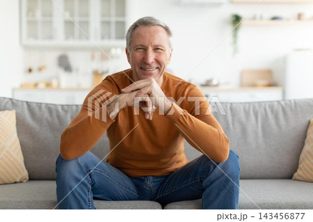 Smiling Mature Caucasian Man Seated Cross-legged On Couch In Cozy Living Room, Exuding Happiness And Contentment, Engaging In Relaxation And Enjoyment At Home. Family Lifestyle Concept. 143456877
