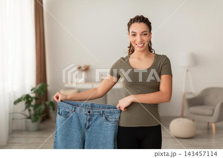 Smiling Young Woman Holding Oversized Jeans, Celebrating Weight Loss Achievement At Home In Modern Living Area. Positive Emotion Reflected As She Shows Progress In Her Fitness Journey. Weight Loss Smiling Young Woman Holding Oversized Jeans, Celebrating Weight Loss Achievement At Home In Modern Living Area. Positive Emotion Reflected As She Shows Progress In Her Fitness Journey. Weight Loss 143457114