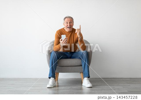 Happy Elderly Caucasian Man Smiling While Using Smartphone And Pointing Up In Living Room At Home. Enjoying Technology And Communication, Excited About Something On Screen. Digital Lifestyle Concept. 143457228