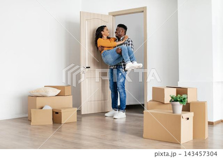 Joyful Emotion. Happy Black Couple In Their 30s Unpacking Boxes While Moving Into New Apartment. Man Lifting Woman With Joyful Expression In Modern Living Room. Moving And Home Concept. Joyful Emotion. Happy Black Couple In Their 30s Unpacking Boxes While Moving Into New Apartment. Man Lifting Woman With Joyful Expression In Modern Living Room. Moving And Home Concept. 143457304