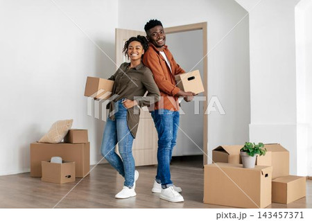 Smiling African American Young Couple Together Holding Moving Boxes In Modern Living Room, Ready For New Home Experience. Joyful Moment Captured In Bright Indoor Setting, Home And Lifestyle Concept. 143457371
