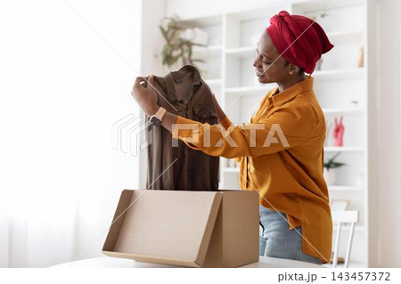 Excited Smiling African American Woman Unboxing New Clothes With Joy In Home Office Setting, Holding A Brown Shirt While Standing By The Table. Unboxing And Shopping Concept. Excited Smiling African American Woman Unboxing New Clothes With Joy In Home Office Setting, Holding A Brown Shirt While Standing By The Table. Unboxing And Shopping Concept. 143457372