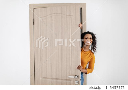 Smiling Young Black Woman Joyfully Peeking Through An Open Door At Home, Creating A Welcoming Atmosphere In A Cozy Interior Setting. Cheerful Emotion Reflected In Her Face. 143457373